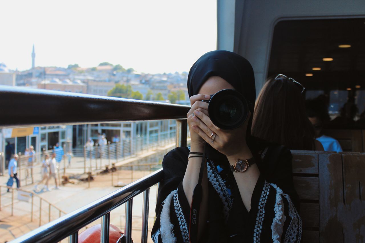 Woman in headscarf photographing cityscape from train with Canon EOS camera