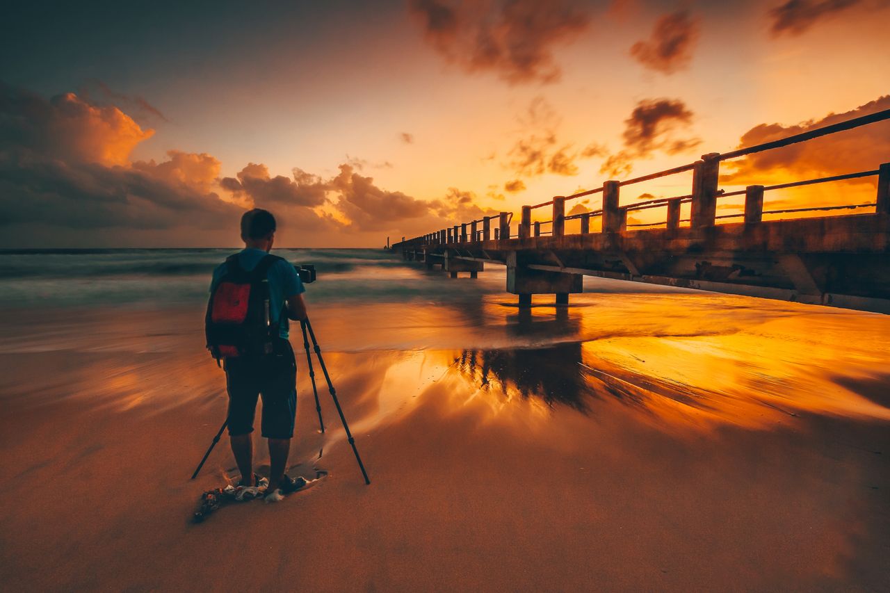 A man with a tripod photographing a stunning sunset by the ocean bridge.