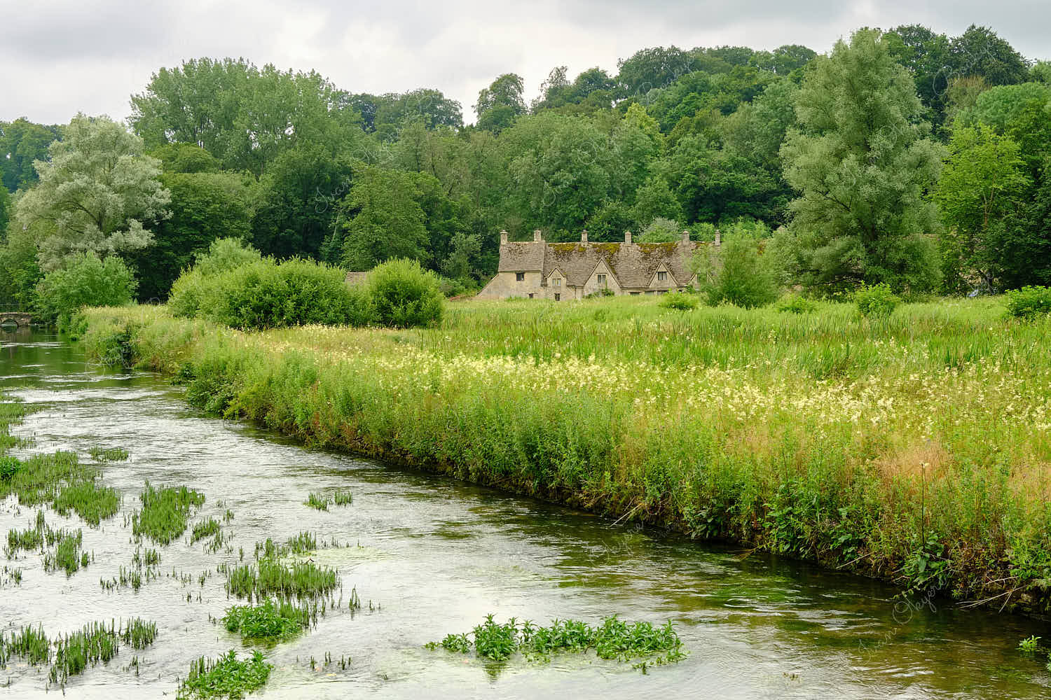 View across River Coln and Rack Isle water meadow to the picturesque Arlington Row cottages, Bibury, Cotswolds, England, UK